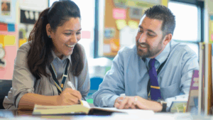 Two smiling teachers seated at a desk, looking at a textbook.